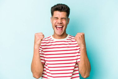 Kafkasyalı genç adam mavi arka planda izole edilmiş kaygısız ve heyecanlı tezahüratlar yapıyor. Zafer konsepti.