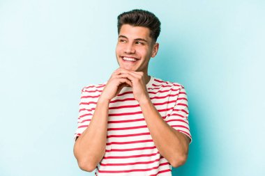 Mavi arka planda izole edilmiş genç beyaz adam ellerini çenesinin altında tutuyor, mutlu bir şekilde yana bakıyor..