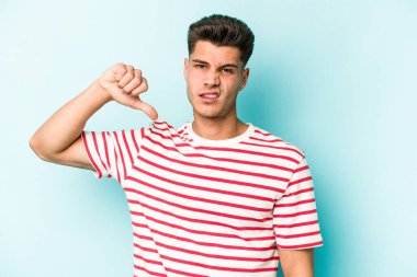 Mavi arka planda izole genç beyaz adam baş parmağını aşağı gösteriyor, hayal kırıklığı konsepti.