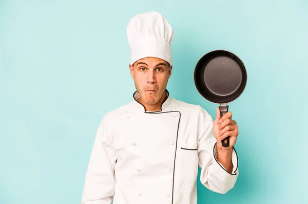 年轻的高加索厨师 拿着被蓝色背景隔离的飞盘 耸了耸肩 睁开眼睛迷惑不解 — 图库照片