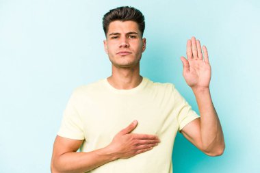 Kafkasyalı genç adam mavi arka planda izole edilmiş yemin ediyor, elini göğsüne koyuyor..