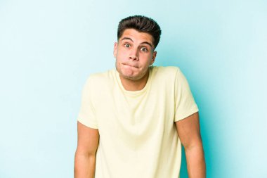 Mavi arka planda izole edilmiş genç beyaz adam omuzlarını silkeliyor ve gözleri açık..