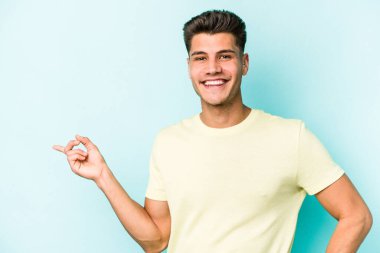 Mavi arka planda izole edilmiş genç beyaz adam işaret parmağıyla işaret ederek gülümsüyor..