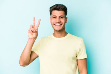 Mavi arka planda izole edilmiş genç beyaz adam zafer işareti gösteriyor ve gülümsüyor..