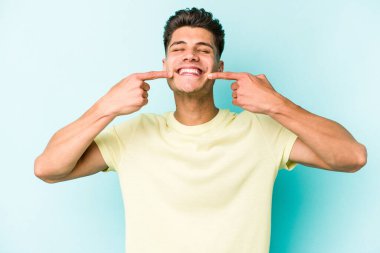Mavi arka planda izole edilmiş genç beyaz adam gülümsüyor, parmaklarını ağıza doğrultuyor..