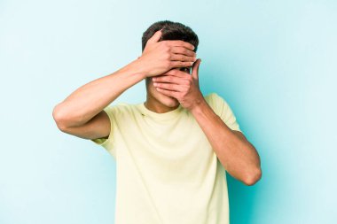 Mavi arka planda izole edilmiş genç beyaz adam kameraya parmaklarıyla göz kırpıyor, yüzünü kapatmaktan utanıyor..