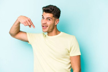 Mavi arka planda izole edilmiş genç beyaz adam bir gömlek fotokopi alanını işaret ediyor, gururlu ve kendinden emin.