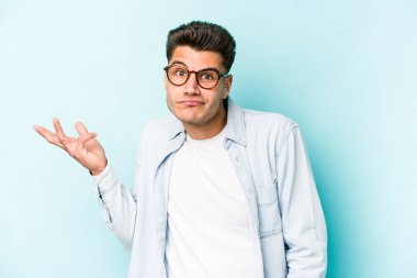 Kafkasyalı genç adam mavi arka planda izole edilmiş sorgulama jestinde şüphe ediyor ve omuz silkiyor..
