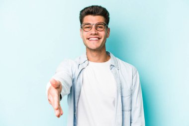 Mavi arka planda izole edilmiş genç beyaz adam selamlama jesti olarak kameraya elini uzatıyor..