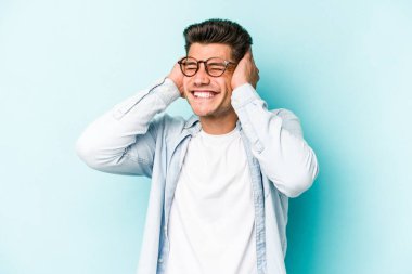 Mavi arka planda izole edilmiş genç beyaz adam kulaklarını elleriyle kapatıyor..