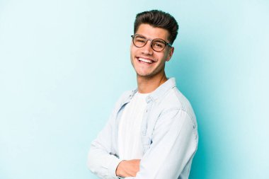 Mavi arka planda kendini güvende hisseden, kararlılıkla kollarını kavuşturan genç beyaz bir adam..