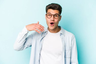Mavi arka planda izole edilmiş genç beyaz bir adam bir şeye gülüyor, ağızlarını ellerle kapatıyor..