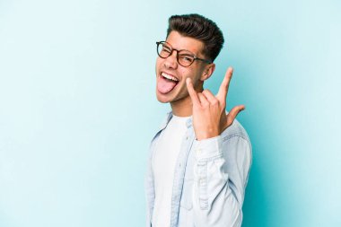 Mavi arka planda izole edilmiş genç beyaz adam parmaklarıyla kaya hareketi gösteriyor.