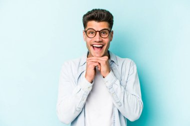 Kafkasyalı genç adam mavi arka planda izole edilmiş şans için dua ediyor, şaşırmış ve ağzını açıp önüne bakıyor..