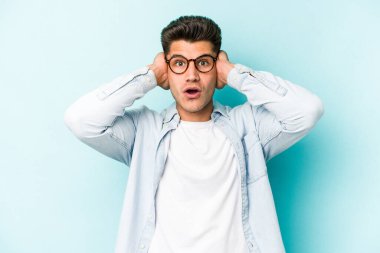 Kafkasyalı genç adam mavi arka planda izole edilmiş kulakları kulaklarıyla kapatıyor çok fazla ses duymamaya çalışıyor..