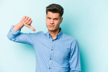 Mavi arka planda izole genç beyaz adam baş parmağını aşağı gösteriyor, hayal kırıklığı konsepti.