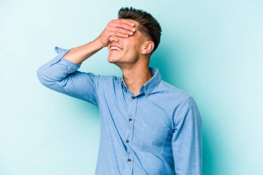 Mavi arka planda soyutlanmış genç beyaz bir adam elleriyle gözlerini kapatıyor, geniş bir gülümsemeyle bir sürpriz bekliyor..