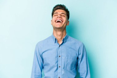 Kafkasyalı genç adam mavi arka planda izole edilmiş rahat ve mutlu bir şekilde gülüyor, boynunda dişlerini gösteriyor..