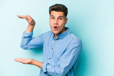 Mavi arka planda izole genç beyaz adam iki eliyle bir şey tutuyor, ürün sunumu..