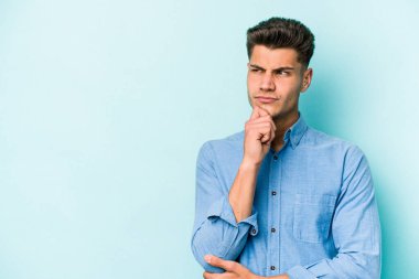 Mavi arka planda izole edilmiş genç beyaz adam şüpheli ve şüpheci bir ifadeyle yana bakıyor..