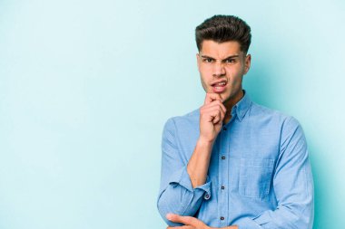 Mavi arka planda izole edilmiş genç beyaz adam şüpheli ve şüpheci bir ifadeyle yana bakıyor..