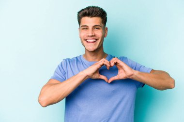 Mavi arka planda izole edilmiş genç beyaz adam gülümsüyor ve elleriyle kalp şeklini gösteriyor..