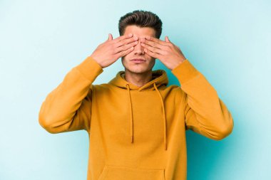 Mavi arka planda izole edilmiş genç beyaz adam elleriyle gözlerini kapatmaktan korkuyor..