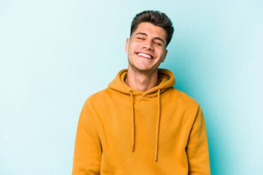 Mavi arka planda izole edilmiş genç beyaz adam gülüyor ve gözlerini kapıyor, rahatlamış ve mutlu hissediyor..