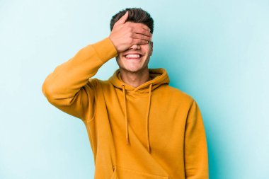 Mavi arka planda soyutlanmış genç beyaz bir adam elleriyle gözlerini kapatıyor, geniş bir gülümsemeyle bir sürpriz bekliyor..
