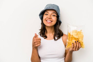 Beyaz arka planda izole edilmiş bir paket cipsi tutan genç İspanyol kadın gülümsüyor ve başparmağını kaldırıyor.