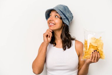 Beyaz arka planda izole edilmiş bir çanta dolusu cipsi tutan genç İspanyol kadın şüpheli ve şüpheci bir ifadeyle yana bakıyor..