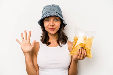 Beyaz arka planda izole edilmiş bir paket cipsi tutan genç İspanyol kadın gülümseyerek beş numarayı parmaklarıyla gösteriyor..