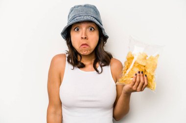 Beyaz arka planda izole edilmiş bir paket cipsi tutan genç İspanyol kadın omuzlarını silker ve gözleri açık kalır..