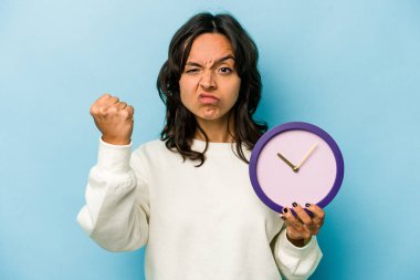 Mavi arka planda bir saat tutan genç İspanyol kadın kameraya yumruk gösteriyor, agresif yüz ifadesi..