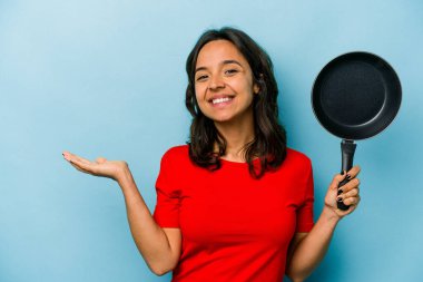 Mavi arka planda izole edilmiş bir tavayı tutan genç İspanyol kadın bir avuç içinin fotokopisini çekiyor ve başka bir elini belinde tutuyor..