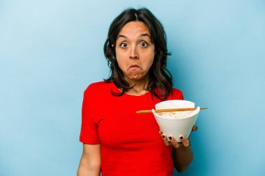 Mavi arka planda izole edilmiş erişte yiyen genç İspanyol kadın omuzlarını silker ve gözleri açık kalır..
