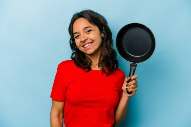 Mavi arka planda izole edilmiş bir tavayı tutan genç İspanyol kadın mutlu, güler yüzlü ve neşeli.