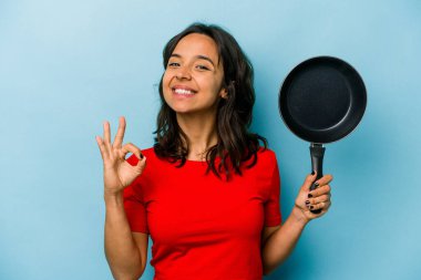 Genç İspanyol kadın elinde bir tava tutuyor mavi arka planda izole edilmiş neşeli ve kendinden emin..