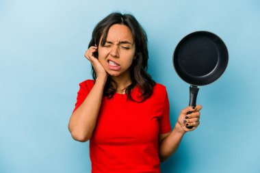 Genç İspanyol kadın elinde tavayla mavi arka planda izole edilmiş kulakları elleriyle kapatıyor..