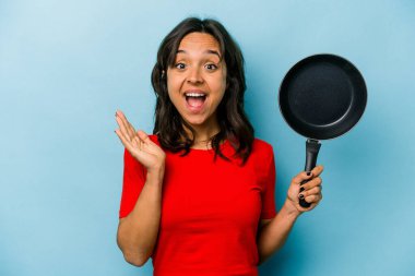 Mavi arka planda izole edilmiş bir tavayı tutan genç İspanyol kadın şaşırdı ve şok oldu..