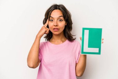 Genç bir İspanyol kadının elinde acemi sürücü işareti var. Beyaz arka planda izole edilmiş. Şakağı işaret ediyor, düşünüyor, bir göreve odaklanıyor..