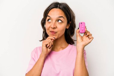 Beyaz arka planda izole edilmiş, şüpheli ve şüpheci bir ifadeyle yan bakan genç İspanyol kadın arabanın anahtarlarını tutuyor..