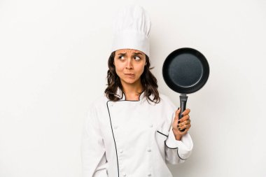 Beyaz arka planda izole edilmiş genç İspanyol aşçı kadın kafası karışmış, şüpheli ve güvensiz hissediyor..