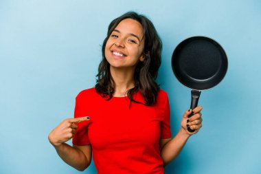 Mavi arka planda izole edilmiş bir tavayı tutan genç İspanyol kadın gömlek fotokopi alanını işaret ediyor, gururlu ve kendinden emin.