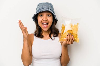 Beyaz arka planda izole edilmiş bir paket cipsi tutan genç İspanyol kadın hoş bir sürpriz alıyor, heyecanlı ve el kaldırıyor..