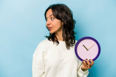 Mavi arka planda bir saat tutan genç İspanyol kadın gülümseyen, neşeli ve keyifli bir kenara bakıyor..