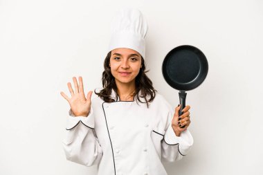 Beyaz arka planda izole edilmiş genç İspanyol aşçı kadın gülümseyerek beş numarayı parmaklarıyla gösteriyor..