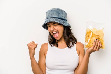 Beyaz arka planda izole edilmiş bir çanta dolusu cipsi tutan genç İspanyol kadın zaferden sonra yumruğunu kaldırıyor, kazanan kavram..