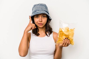 Beyaz arka planda izole edilmiş bir paket cipsi tutan genç İspanyol kadın şakağı parmağıyla işaret ediyor, düşünüyor ve bir göreve odaklanıyor..