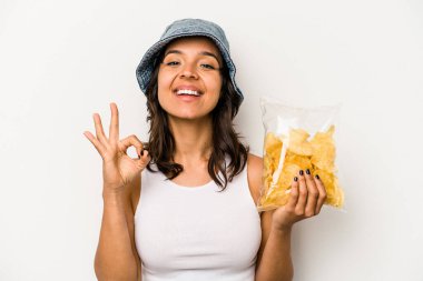 Beyaz arka planda izole edilmiş bir paket cipsi tutan genç İspanyol kadın neşeli ve kendinden emin bir şekilde iyi niyet gösteriyor..